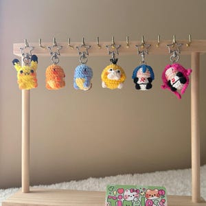 May include: A wooden display stand holds seven colorful, crocheted character keychains. The keychains feature various designs in yellow, orange, blue, and pink. Each keychain hangs from a silver star-shaped hook. A small card with the text "Timeless Kitty" is at the base.