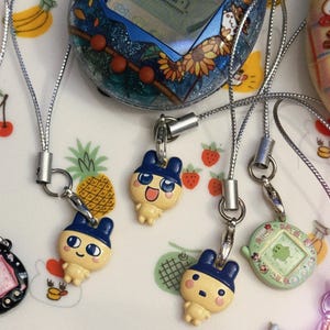 May include: A collection of vintage handheld digital pets, including a blue and glittery Tamagotchi with a sunflower design, and several character charms with silver lanyards. The background is a white plate with fruit and animal illustrations.