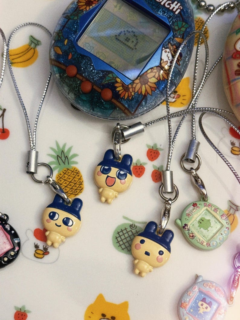 May include: A collection of vintage handheld digital pets, including a blue and glittery Tamagotchi with a sunflower design, and several character charms with silver lanyards. The background is a white plate with fruit and animal illustrations.
