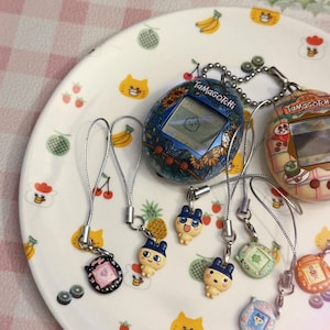 May include: A collection of vintage Tamagotchi digital pet toys and charms. The handheld devices feature screens and buttons, with various designs including floral patterns and cartoon characters. The charms are attached to silver cords.