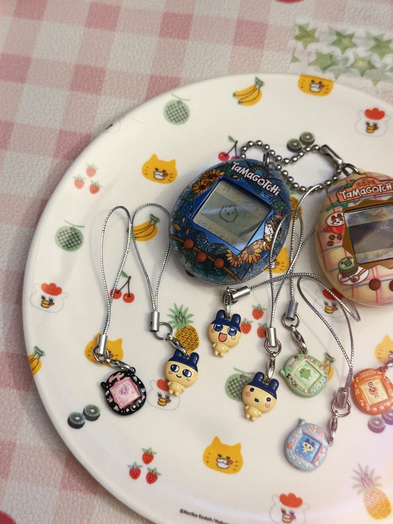 May include: A collection of vintage Tamagotchi digital pet toys and charms. The handheld devices feature screens and buttons, with various designs including floral patterns and cartoon characters. The charms are attached to silver cords.