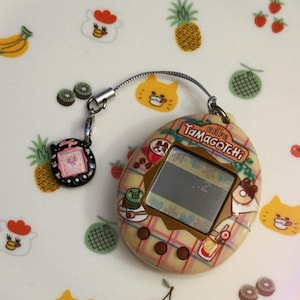 May include: A vintage Tamagotchi Cafe electronic pet game with a plaid design, featuring a small screen and a connected charm. The background is decorated with fruit and animal illustrations, including cats, pineapples, and cherries.
