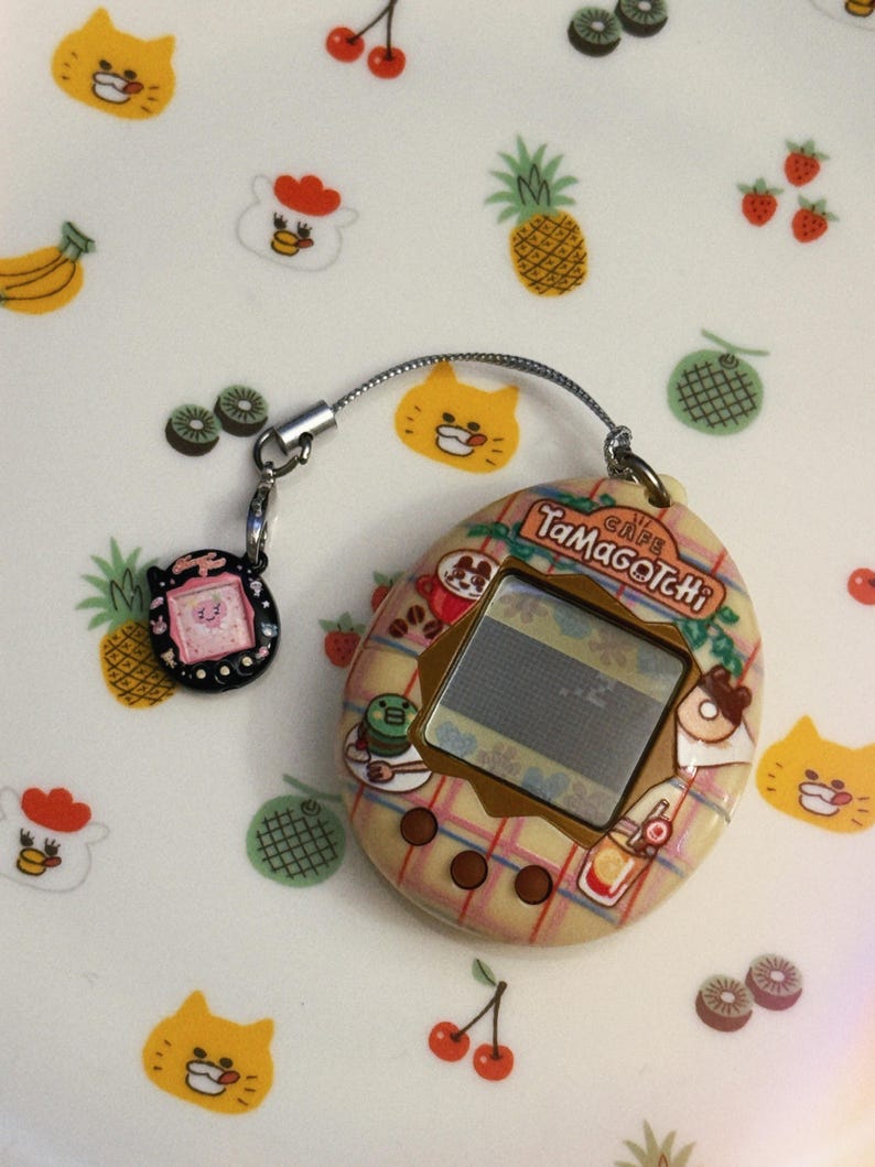 May include: A vintage Tamagotchi Cafe electronic pet game with a plaid design, featuring a small screen and a connected charm. The background is decorated with fruit and animal illustrations, including cats, pineapples, and cherries.