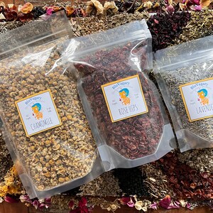 Pode incluir: Tr&ecirc;s sacos de pl&aacute;stico transparentes cheios de ervas e flores secas. Os sacos s&atilde;o etiquetados com "Camomila", "Rosa Mosqueta" e "Lavanda". Os sacos est&atilde;o sobre uma superf&iacute;cie de madeira com flores e ervas secas espalhadas ao redor.