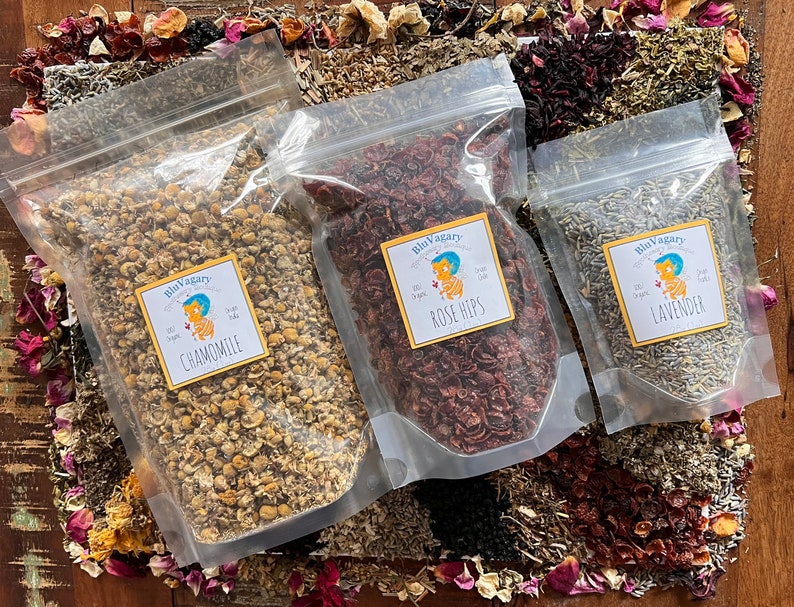 Pode incluir: Tr&ecirc;s sacos de pl&aacute;stico transparentes cheios de ervas e flores secas. Os sacos s&atilde;o etiquetados com "Camomila", "Rosa Mosqueta" e "Lavanda". Os sacos est&atilde;o sobre uma superf&iacute;cie de madeira com flores e ervas secas espalhadas ao redor.