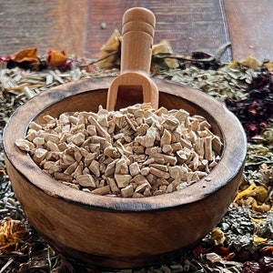 Pode incluir: Uma tigela de madeira cheia de peda&ccedil;os de raiz de bardana secos, de cor castanho claro e irregulares. Uma concha de madeira est&aacute; apoiada na borda da tigela.