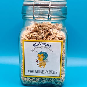 Pode incluir: Um frasco de vidro cheio de peda&ccedil;os secos de raiz de ang&eacute;lica, de cor castanho claro e forma irregular. O frasco tem um fecho de metal e um r&oacute;tulo amarelo com o texto "Blu Vagary Apothecary Boutique...where wellness wanders..." e uma ilustra&ccedil;&atilde;o de abelha de desenho animado.