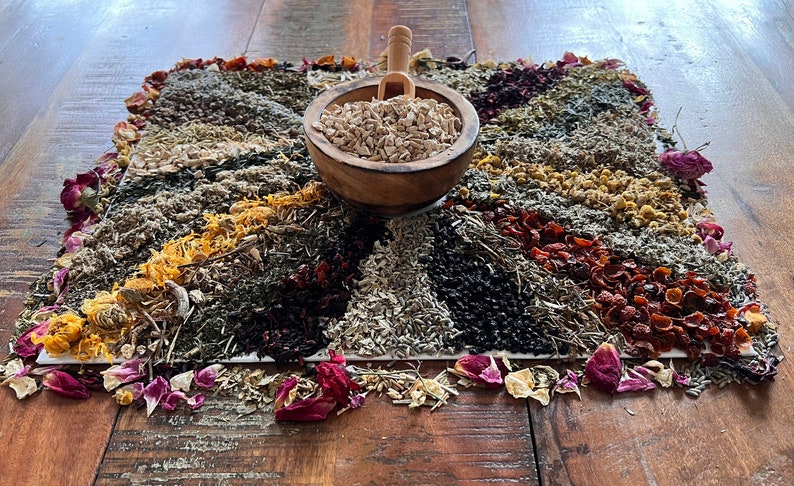 Pode incluir: Uma variedade de ervas e flores secas dispostas em um padr&atilde;o circular sobre uma superf&iacute;cie de madeira. As ervas s&atilde;o de v&aacute;rias cores, incluindo vermelho, laranja, amarelo, verde e marrom. Uma tigela de madeira cheia de ervas secas est&aacute; no centro da disposi&ccedil;&atilde;o.