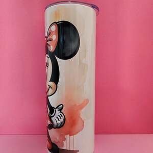 May include: A white stainless steel tumbler with a watercolor design of Minnie Mouse. Minnie is wearing a red dress with white polka dots and a red bow. The background is a light orange and yellow watercolor wash.