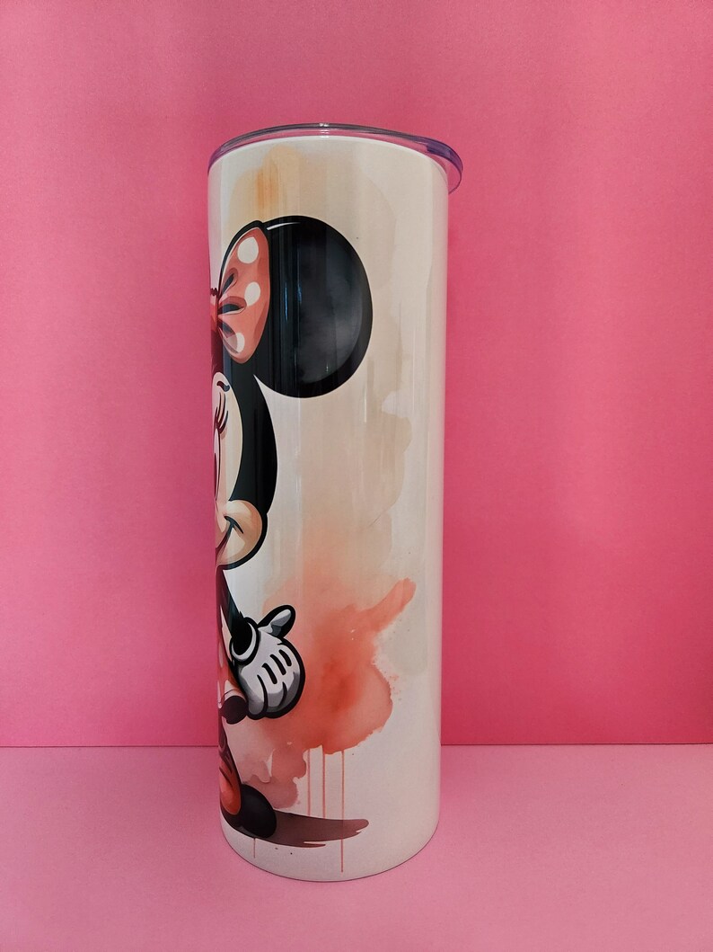 May include: A white stainless steel tumbler with a watercolor design of Minnie Mouse. Minnie is wearing a red dress with white polka dots and a red bow. The background is a light orange and yellow watercolor wash.