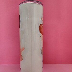 May include: A white stainless steel tumbler with a lid. The tumbler has a design of abstract watercolor-like shapes in shades of pink, orange, and brown.