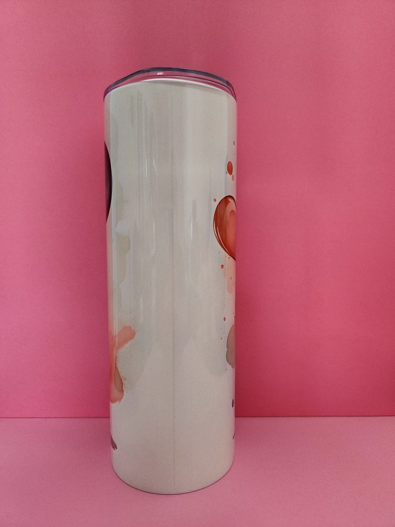 May include: A white stainless steel tumbler with a lid. The tumbler has a design of abstract watercolor-like shapes in shades of pink, orange, and brown.