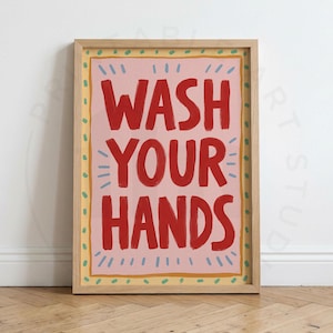 Pode incluir: Uma impressão de arte emoldurada com o texto WASH YOUR HANDS em letras vermelhas em negrito sobre um fundo rosa claro. O design inclui uma borda amarela com traços verdes e marcas de destaque azul claro ao redor do texto central.