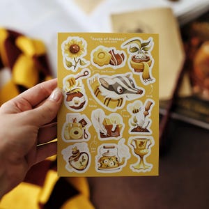 May include: A sheet of yellow stickers featuring various illustrations, including a sunflower, honey pot, badger, camera, and teapot. The text "house of kindness" is at the top. The stickers are held by a hand.