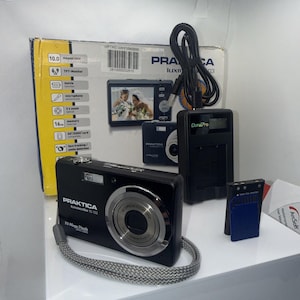 May include: A black Praktica Luxmedia 10-03 digital camera with a silver lens, a grey wrist strap, a black battery charger with a cord, a battery, and a blue memory card. The camera is displayed in front of its original yellow and white box.