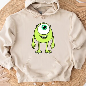 Puede incluir: Sudadera con capucha beige con un personaje de dibujos animados verde lima en la parte delantera. El personaje tiene un ojo grande, una boca sonriente y brazos y piernas peque&ntilde;os. La sudadera tiene un bolsillo de canguro.