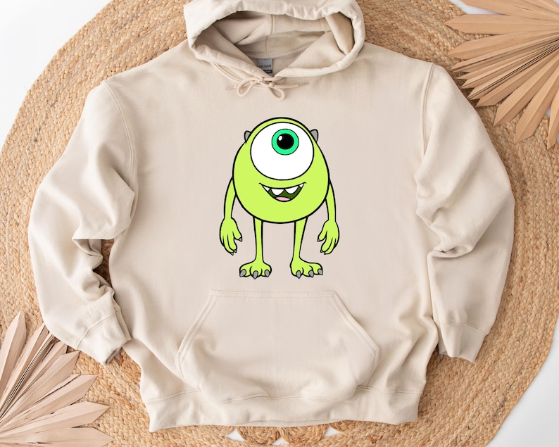 Puede incluir: Sudadera con capucha beige con un personaje de dibujos animados verde lima en la parte delantera. El personaje tiene un ojo grande, una boca sonriente y brazos y piernas peque&ntilde;os. La sudadera tiene un bolsillo de canguro.