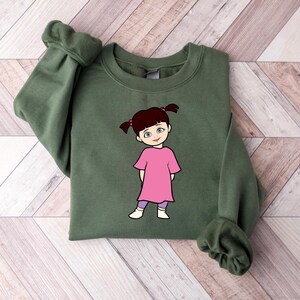 Puede incluir: Una sudadera verde oliva con una ilustraci&oacute;n de dibujos animados de una ni&ntilde;a con un vestido rosa y leggings morados. La ni&ntilde;a tiene coletas marrones y est&aacute; sonriendo. La sudadera tiene mangas largas con pu&ntilde;os acanalados.