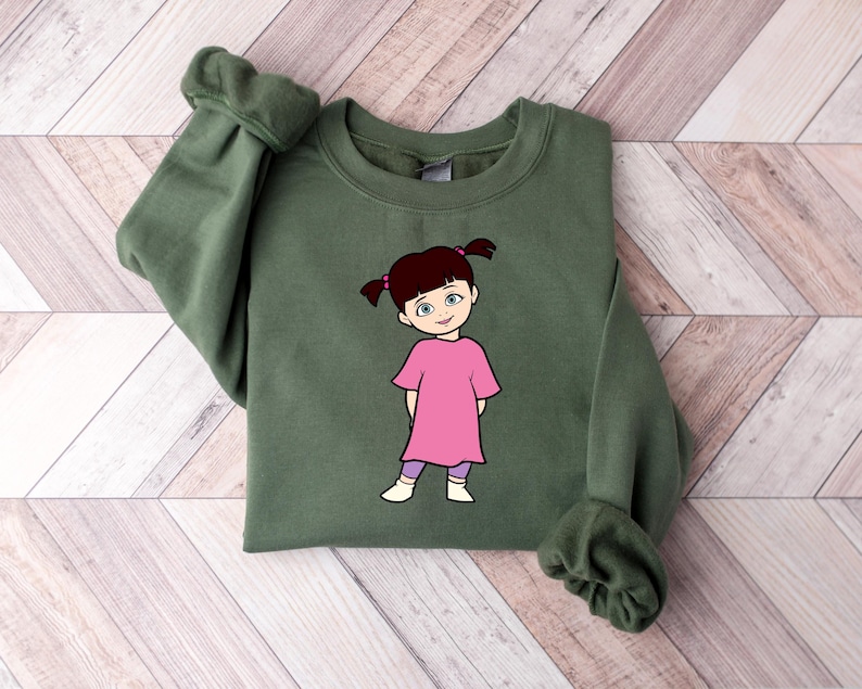 Puede incluir: Una sudadera verde oliva con una ilustraci&oacute;n de dibujos animados de una ni&ntilde;a con un vestido rosa y leggings morados. La ni&ntilde;a tiene coletas marrones y est&aacute; sonriendo. La sudadera tiene mangas largas con pu&ntilde;os acanalados.