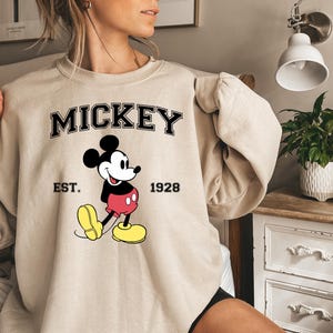 May include: Beige sweatshirt featuring a graphic of Mickey Mouse. The word "MICKEY" is printed in black block letters above the cartoon character. Below the word "MICKEY" is the text "EST. 1928". Mickey Mouse is wearing red shorts and yellow shoes.