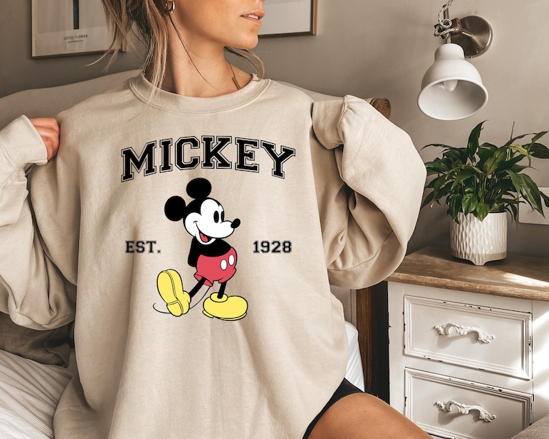 May include: Beige sweatshirt featuring a graphic of Mickey Mouse. The word "MICKEY" is printed in black block letters above the cartoon character. Below the word "MICKEY" is the text "EST. 1928". Mickey Mouse is wearing red shorts and yellow shoes.