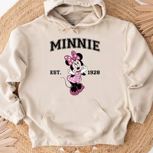 May include: A beige hooded sweatshirt featuring a Minnie Mouse graphic. The graphic includes the word "MINNIE" in black block letters above an image of Minnie Mouse in a pink polka-dot dress. The text "EST. 1928" is below the graphic.