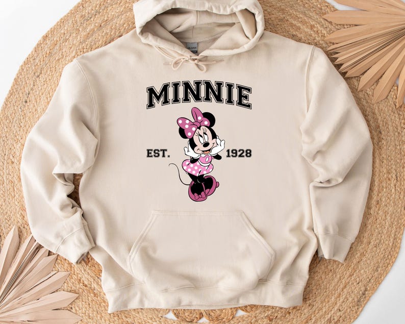 May include: A beige hooded sweatshirt featuring a Minnie Mouse graphic. The graphic includes the word "MINNIE" in black block letters above an image of Minnie Mouse in a pink polka-dot dress. The text "EST. 1928" is below the graphic.