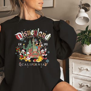 Pode incluir: Camisola preta com um gr&aacute;fico da Disneyland com Mickey Mouse, Pato Donald e outros personagens. O design inclui um castelo, flocos de neve e o texto "Disneyland Est. 1955 California."
