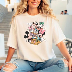 May include: White t-shirt with a colorful graphic print of Mickey Mouse, Minnie Mouse, Donald Duck, Goofy, and Pluto. The characters are arranged in a cluster, with Mickey Mouse and Minnie Mouse in the front.