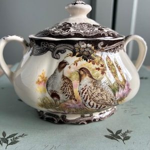 May include: A cream-coloured ceramic sugar bowl with a lid. The lid and rim feature a brown floral and lace pattern. The bowl has two handles and a painted scene of two birds in a field.