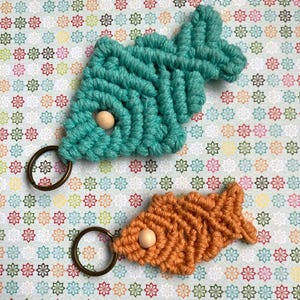 May include: Two macrame fish keychains with wooden beads. The fish are teal and orange. The keychains are on a white background with a floral pattern.