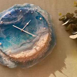 May include: A round wall clock with a resin design resembling an ocean scene. The clock face features shades of blue and white, with white accents. The clock hands are silver. The clock is a decorative item.