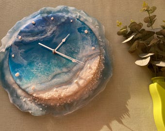 May include: A round wall clock with a resin design resembling an ocean scene. The clock face features shades of blue and white, with white accents. The clock hands are silver. The clock is a decorative item.