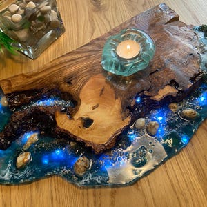 Peut inclure: Un plateau de service en bois avec une rivière en résine bleue qui le traverse. La rivière est remplie de petites pierres blanches et comporte un bougeoir en verre turquoise au centre. Le plateau est parfait pour servir des collations ou des boissons.