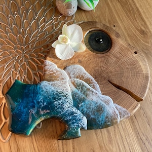 May include: A wooden serving board shaped like a fish with a blue and white resin ocean wave design. The board has a small hole for hanging and a round cutout for serving.