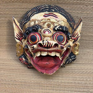 Puede incluir: Una máscara de madera tallada a mano que presenta un rostro estilizado con grandes ojos saltones, una boca muy abierta con dientes visibles y orejas puntiagudas. La máscara está pintada en tonos crema, negro, rojo y dorado con texturas.