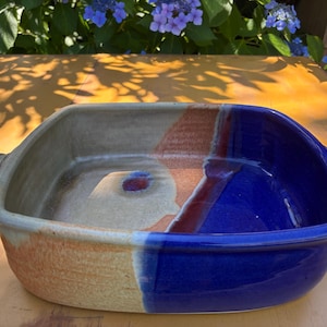 Puede incluir: Una fuente de horno cuadrada de cerámica con esquinas redondeadas y dos pequeñas asas laterales. El interior y el exterior presentan un diseño bicolor, con una mitad en azul cobalto brillante y la otra en beige mate con un acento marrón rojizo.
