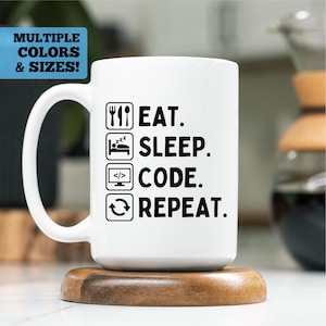 Peut inclure: Mug en céramique blanche avec le texte "EAT. SLEEP. CODE. REPEAT." en noir, ainsi que des icônes associées. Le mug est posé sur un dessous de verre en bois. Le coin supérieur gauche de l'image contient le texte "MULTIPLE COLORS & SIZES!"