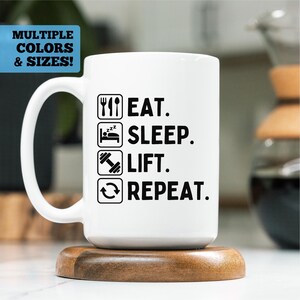 May include: White ceramic mug with the text "EAT. SLEEP. LIFT. REPEAT." in black. The mug also features icons of a fork and knife, a bed, a dumbbell, and a recycling symbol. The mug is on a wooden coaster.