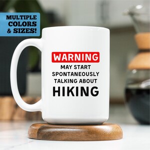 May include: White ceramic mug with a large handle, featuring the text "WARNING MAY START SPONTANEOUSLY TALKING ABOUT HIKING" in black and red. The mug sits on a wooden coaster. The top left corner has a blue rectangle with the text "MULTIPLE COLORS & SIZES!"