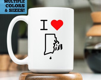 May include: White ceramic coffee mug with a black outline of the state of Rhode Island and the text "I love" with a red heart.