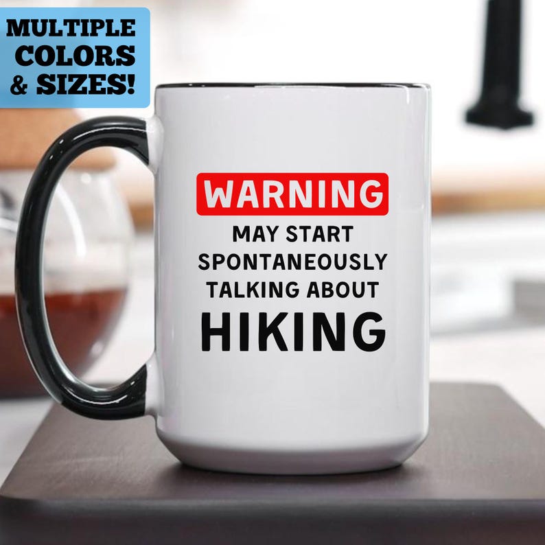 May include: White ceramic mug with a black handle and rim. The mug has the text "WARNING MAY START SPONTANEOUSLY TALKING ABOUT HIKING" in black, with "WARNING" in a red rectangle. A blue rectangle in the top left corner reads "MULTIPLE COLORS & SIZES!"