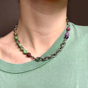 Op de afbeelding: Een zilveren ketting met groene en paarse kralen. De ketting is verbonden met een rij groene en rode kralen.