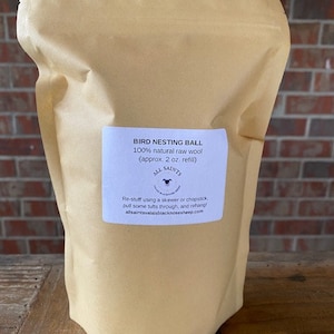 May include: A brown paper bag filled with a bird nesting ball made of 100% natural raw wool. The bag has a label that reads "Bird Nesting Ball 100% natural raw wool (approx. 2 oz. refill) All Saints Re-stuff using a skewer or chopstick, pull some tufts through, and rehang Allsaintsvalaisblacknosesheep.com"