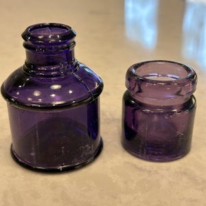 May include: Two antique, deep purple glass inkwells. The larger inkwell has a rounded body and a flared rim. The smaller inkwell is cylindrical with a wide opening. Both are made of a dark, translucent glass.