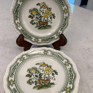 May include: Two white ceramic plates with scalloped edges, featuring a central floral motif in yellow, orange, and green. The border is decorated with a green and yellow geometric pattern. One plate is displayed upright on a dark wooden stand.