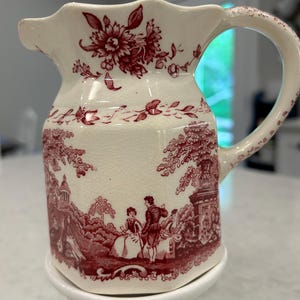 May include: Cream-colored ceramic pitcher with a red floral and pastoral scene design. The pitcher features a scalloped rim, a curved handle, and a detailed illustration of people in a landscape. The design is repeated around the body of the pitcher.
