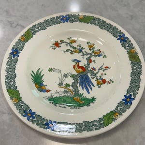 May include: A white ceramic plate with a colorful bird and tree design. The plate features a border of green leaves and blue flowers. The central image depicts a bird with red, blue, and yellow plumage perched on a tree with colorful blossoms.