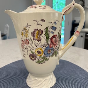 May include: Cream-colored ceramic pitcher with a lid, adorned with a floral pattern in shades of purple, yellow, red, and blue. The pitcher has a curved handle and a spout. The base has a decorative, ridged design.
