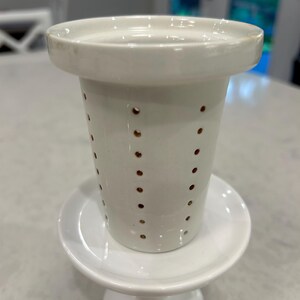 May include: A white ceramic tea infuser with a wide rim and a tapered body featuring vertical rows of small, evenly spaced circular perforations. The infuser is resting on a matching white ceramic saucer with a pedestal base.